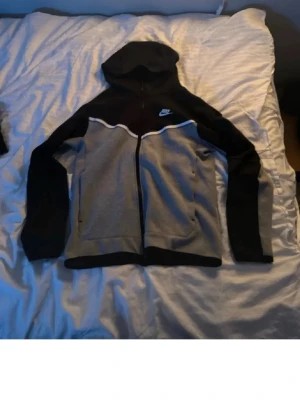 Nike tech grå/svart - svart grå Nike tech som är köpt för ett år sen ungefär. De enda är ett litet hål vid sidan av dragkedjan längst ner på hoodien men inget som syns super mycket.