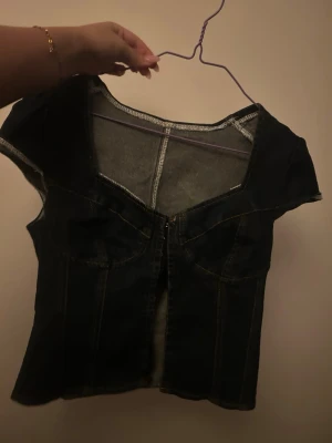 Mörkblå denim korsett-topp - Snygg mörkblå korsett-topp i denim med markerade sömmar och hakknäppning framtill. Toppen har korta ärmar och en figurnära passform som framhäver siluetten. Perfekt att styla med jeans eller kjol för en cool look.