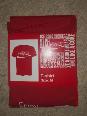 Röd Coca-Cola t-shirt i bomull - Säljer en röd t-shirt från Coca-Cola med klassiskt vitt tryck framtill. T-shirten är kortärmad och har en normal passform. Tillverkad i 100% bomull för en skön känsla. Perfekt för dig som gillar retro och ikoniska loggor.