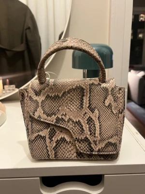 ATP Montalcino handväska i ormmönstrat skinn - Säljer en snygg beige handväska med ormmönster och stilren form. Väskan har ett topphandtag och avtagbar axelrem, perfekt för att bära på olika sätt. Materialet är skinn med struktur och väskan stängs med lock. Riktigt trendig accessoar för dig som gillar statement-detaljer.