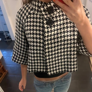Svartvit rutig croppad jacka - Säljer en croppad jacka i svartvitt hundtandsmönster med stora knappar framtill och trekvartsärmar. Jackan har en boxig passform och är perfekt för dig som vill sticka ut. Materialet känns som en blandning av ull och syntet. Snygg till både jeans och kjol.🖤🤍
