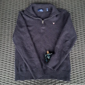 Mörkgrå half zip tröja från GANT - Snygg Marinblå stickad tröja från GANT med broderad logga på bröstet. Tröjan har half zip-dragkedja och hög krage, perfekt för lager-på-lager. Långärmad och i mjukt material som håller dig varm under kyliga dagar. Tröjan ser ut som att vara grå på bilden men den är mer än marinblå färg