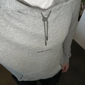 Grå Saint Laurent hoodie med snörning - Säljer en grå hoodie från Saint Laurent med minimalistisk logga på bröstet. Tröjan har huva med snörning, känguruficka och långa ärmar. Perfekt för dig som gillar stilren streetwear och vill ha något bekvämt och snyggt. ( Strl S) personen i Bild 1,83m