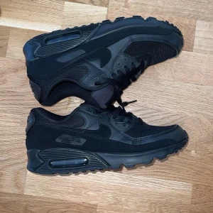 Nike Air Max 90 Triple Black 42 - Säljer ett par helsvarta Nike Air Max 90 med klassisk chunky sula och synlig Air-enhet. Skorna har mesh och syntetpaneler, snörning och diskret Nike-logga på sidan. Perfekta för dig som gillar en clean och sportig look.