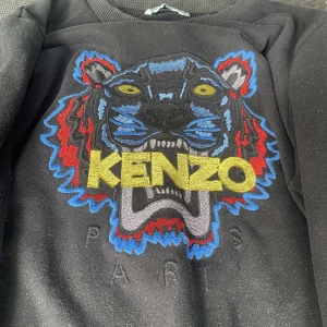 Svart Kenzo sweatshirt med tigerbrodyr - Svart sweatshirt från Kenzo med ikonisk tigerbrodyr i blått, rött, gult och grått framtill. Stort KENZO-tryck i gult över bröstet och Paris-brodyr under. Klassisk rund halsringning och långa ärmar. Perfekt statement-plagg för dig som gillar streetstyle.