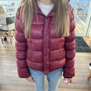 Vinröd pufferjacka  - Snygg vinröd pufferjacka från Superdry med glansig finish och huva. Jackan har breda, quiltade sektioner, dragkedja framtill och två stora fickor. Perfekt för kalla dagar och ger en cool vibe till din outfit. Trendig och går inte att köpa längre. Storlek 36/S, TRYCK INTE PÅ KÖP NU INNAN DU SKRIVIT TILL MIG 