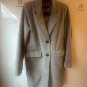 Grå kappa från Vero Moda - Stilren grå kappa från Vero Moda med klassisk krage och två knappar framtill. Jackan har raka linjer, långa ärmar och två fickor med lock. Perfekt för dig som gillar en clean och tidlös look.