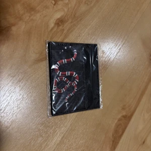 Svart kortfodral med orm  - Snyggt svart kortfodral  med ikoniskt GG-mönster och en röd, vit och svart orm på framsidan. Perfekt för kort och ID, smidig att ha i fickan. Tillverkat i tåligt syntetmaterial med stilren design.
