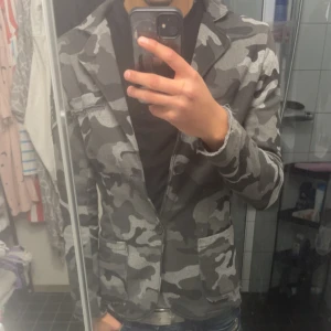 Grå camo kavaj/overshirt - Säljer en snygg kavaj i grått camouflage-mönster. Kavajen har två knappar framtill, klassisk krage och två fickor. Perfekt för dig som vill sticka ut med en unik stil. Passar till både jeans och mer dressade outfits.