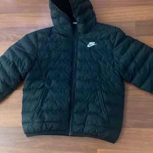 Svart pufferjacka från Nike med huva - Svart pufferjacka från Nike med huva och dragkedja framtill. Jackan har quiltad design och klassisk Nike-logga på bröstet. Perfekt för kalla dagar och riktigt snygg streetstil. Tillverkad i syntetmaterial för extra värme.PRIS KAN DISKUTERAS 