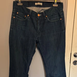 Mörkblå jeans från Acne Jeans - Snygga mörkblå jeans från Acne Jeans med klassisk femficksdesign och orange läderpatch bak. Jeansen har raka ben och subtila slitningar för en cool look. Tillverkade i robust denim med snygga kontrastsömmar och kopparfärgade nitar.