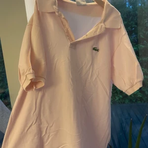 Ljusrosa pikétröja från Lacoste - Säljer en ljusrosa pikétröja från Lacoste med klassisk krage och korta ärmar. Tröjan har den ikoniska gröna krokodilloggan broderad på bröstet och är tillverkad i mjuk bomull. Perfekt för en clean och sportig look.