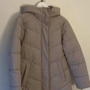 Beige pufferjacka från McKinley, strl 36 - Säljer en beige pufferjacka från McKinley i storlek 36. Jackan har huva, dragkedja och tryckknappar framtill samt två fickor. Perfekt för kalla dagar med sin quiltade design och mjuka material. Enkel och stilren look som passar till det mesta.
