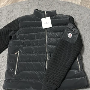 Svart Moncler pufferjacka S - Svart pufferjacka från Moncler med stickade ärmar och quiltad kropp. Jackan har dragkedja framtill, två dragkedjefickor och Moncler-logga på ena ärmen. Materialmix av polyester och stickat ger en modern look. Perfekt för kyliga dagar.