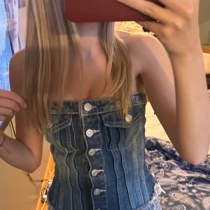 Blå jeans korsett tubtopp - Säljer en snygg blå jeans korsett tubtopp med knappar framtill och coola sömmar. Toppen är axelbandslös och har fickdetaljer upptill. Perfekt för dig som vill ha en unik och trendig denimlook. Passar dig som gillar att sticka ut!