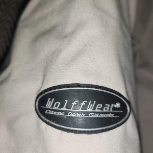 Beige dunjacka från Wolffwear - Snygg beige dunjacka från Wolffwear med quiltat svart foder och hög krage. Jackan har dragkedja, tryckknappar, två fickor framtill och ribbade muddar vid ärmslut och nederkant. Perfekt för kalla dagar och riktigt skön att bära.