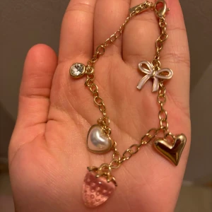 Guldigt armband med berlocker - Charmigt armband i guldfärgad metall med flera söta berlocker: en rosa jordgubbe, ett pärlformat hjärta, ett guldhjärta, en liten rosett och en glittrig sten. Perfekt accessoar för dig som gillar lekfulla detaljer och pastelliga färger.