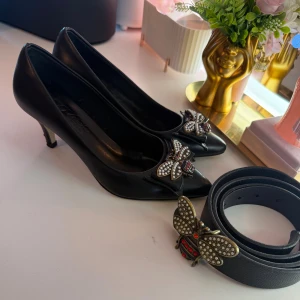 Svarta pumps med pärlbeströdd bi-detalj bält och skor  - Snygga svarta pumps i skinn med spetsig tå och låg klack. Framtill sitter en stor bi-detalj i antikguld med pärlor och röda stenar som ger en unik och lyxig vibe. Perfekt för dig som gillar statement-accessoarer och vill sticka ut.både bält och skor kostar 400 kr