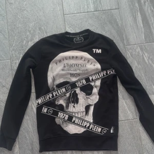 Svart Philipp Plein tröja  - Svart långärmad tröja från Philipp Plein med stor dödskalleprint och logga framtill. Snygga detaljer med textband och tryck även på ryggen. Crewneck-modell i mjuk bomull.