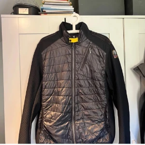 Svart quiltad jacka Parajumpers L - Snygg svart quiltad jacka från Parajumpers i storlek L. Jackan har dragkedja framtill, bröstficka med dragkedja och coola detaljer som gul rem vid kragen. Materialmix med glansig syntet och stickade partier på ärmar och sidor.