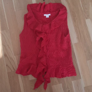 Röd festlig blus från Gina Tricot - Snygg röd blus från Gina Tricot med volangdetaljer längs krage och framkant. Västen har en crinklad struktur och knyt framtill. Perfekt för dig som vill sticka ut med färg och coola detaljer.