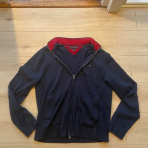 Marinblå kofta från Tommy Hilfiger - Snygg marinblå kofta från Tommy Hilfiger med dragkedja framtill och ribbade muddar. Insidan av kragen är röd och ger en schysst kontrast. Liten broderad logga på bröstet och två fickor framtill. Perfekt för lager-på-lager under höst och vinter.