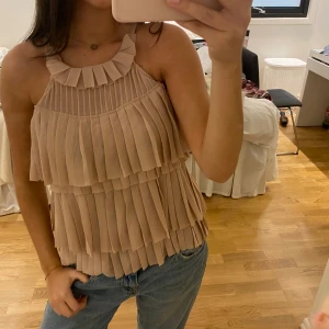 Beige plisserad linnetopp H&M Conscious - Säljer en beige linnetopp från H&M Conscious Exclusive i storlek 34. Toppen har flera lager med plisserade volanger och ett snyggt halterneckband med veckad detalj. Materialet är lätt och luftigt, perfekt för varma dagar. Supersnygg att styla med jeans eller kjol! Rensar garderoben så köp gärna och kom med bud!