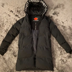 Svart lång pufferjacka Zavetti Canada - Svart lång pufferjacka från Zavetti Canada med stor huva och vit logotyptext runt kanten. Jackan har dragkedja, två fickor med dragkedja och en röd lönnlöv-detalj. Perfekt för kalla dagar och riktigt snygg streetstil.