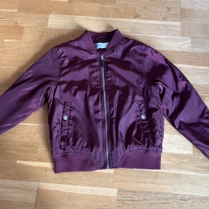 Vinröd bomberjacka med volangdetalj - Snygg vinröd bomberjacka från U i glansigt syntetmaterial. Jackan har dragkedja framtill, ribbade muddar och coola volangdetaljer på ryggen och ärmarna. Två fickor med tryckknappar framtill.