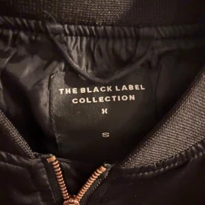 Svart bomberjacka från The Black Label - Snygg svart bomberjacka från The Black Label Collection i storlek S. Jackan har en glansig finish, ribbade muddar och detaljer i guld på dragkedjorna. Klassisk passform med fickor på sidorna och ärmen. Perfekt för dig som vill ha en stilren och trendig look.