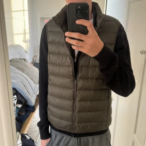 Olivgrön dunväst från Uniqlo S - Säljer en olivgrön dunväst från Uniqlo Ultra Light Down i storlek S. Västen är quiltad med horisontella sömmar, har dragkedja framtill och står för en clean och sportig look. Perfekt att slänga över en hoodie eller sweatshirt när det är kyligt ute.