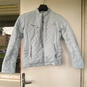 Ljusblå quiltad jacka med dragkedjor - Säljer en ljusblå quiltad jacka med coola detaljer som silverfärgade dragkedjor och spännen i midjan. Jackan har lång ärm, ståkrage och fyra fickor med dragkedja framtill. Perfekt för dig som gillar stilren men ändå unik look.