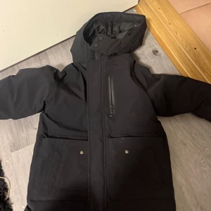 Barn  pufferjacka från ZARA med huva - Svart pufferjacka från ZARA med stor huva, dragkedja och knappar framtill. Jackan har två stora fickor med tryckknappar och en bröstficka med dragkedja. Insidan är quiltad och jackan är perfekt för kalla dagar. 6-7 år men zara är ju lite större i storleken