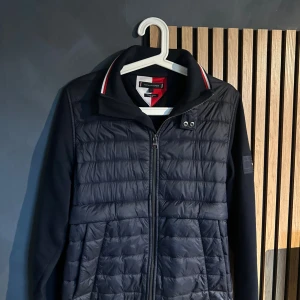 Tommy hilfiger cardigan  - Svart pufferjacka från Tommy Hilfiger med quiltad framsida och slät baksida. Jackan har dragkedja, ståkrage med röd och vit rand samt logotyp på ärmen. Materialmix med polyester och bomull, perfekt för dig som gillar sportig och clean stil. Väldigt fint skick och fet nypris 2500kr mitt pris 999 strl S 