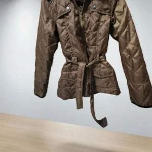 Quiltad brun jacka från b.young - Snygg quiltad jacka från b.young i brun färg med bälte i midjan och dragkedja framtill. Jackan har långa ärmar, tryckknappar på axlar och fickor, samt ett stilrent mönster. Perfekt för dig som gillar en clean look med lite edge.