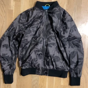 Svart bomberjacka med camomönster från Diesel - Snygg svart bomberjacka från Diesel med grått camouflagemönster. Jackan har dragkedja framtill, ribbade muddar och krage samt två snedställda fickor med dragkedja. Tillverkad i lätt polyester med blått innerfoder för extra edge.