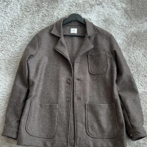 Brun overshirt från John Henric - Säljer en brun overshirt från John Henric i ull. Jackan har tre stora utanpåfickor, klassisk krage och knäppning framtill. Perfekt lager-på-lager-plagg med stilren och enkel design.