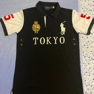 Polo Ralph Lauren Chief Keef Tokyo - Snygg svart och vit pikétröja från Polo Ralph Lauren med broderad Tokyo-text, guldigt emblem och stor vit polospelare på bröstet. Röda siffror på ärmar och rygg. Slim fit-modell i 100% bomull. Perfekt för dig som gillar sportig och stilren look.