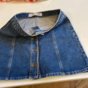 Blå jeans minikjol med knappar framtill - Snygg blå jeans minikjol från Denim Co. med A-linjeform och dekorativa sömmar. Kjolen har knappar hela vägen framtill och klassisk midjehöjd. Perfekt till sneakers eller boots för en cool look. Liten i storlek passar också dig med storlek 38