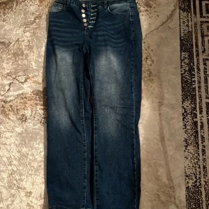 Mörkblå jeans med hög midja och knappar - Snygga mörkblå jeans med hög midja och flera silverfärgade knappar framtill. Jeansen har raka ben, klassiska fickor fram och bak samt en lätt tvättad look över låren. Perfekta för dig som gillar en trendig och bekväm stil.