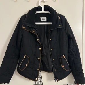 Svart quiltad jacka från Vero Moda - Svart quiltad jacka med guldiga knappar och dragkedjor i storlek S från Vero Moda. Jackan har bruna detaljer längs kanterna, hög krage och två fickor framtill med dragkedja. Jackan är i bra skick. 