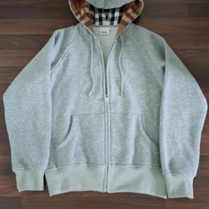 Grå hoodie från Burberry med dragkedja - Snygg grå hoodie från Burberry med klassiskt rutigt foder i huvan, broderad logga på bröstet och dragkedja framtill. Tröjan har snörning i huvan och två fickor. Perfekt för en chill och stilren look. Sizeb är mellan s och m och du får med Burberry påse och prislapp.