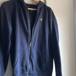 Marinblå hoodie från Lyle & Scott - Snygg marinblå hoodie från Lyle & Scott med gul logga på bröstet. Tröjan har huva, dragkedja och två stora fickor framtill. Perfekt för chill dagar och enkel att matcha med jeans eller joggers.storlek 14/15år/S