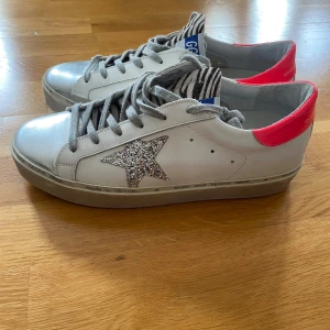 Golden Goose vita sneakers med glitterstjärna - Säljer ett par vita Golden Goose sneakers med grå snörning, silverglittrig stjärna på sidan och rosa detalj vid hälen. Skorna har en klassisk låg siluett och randiga detaljer på plösen. Modell Hi star limited edition. Nypris dryga 6000kr. Endast använda några enstaka gånger. Perfekta för dig som vill sticka ut med en cool och trendig look.