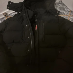 Svart pufferjacka med avtagbar fuskpäls - Svart pufferjacka med avtagbar fuskpäls på huvan. Jackan har dragkedja framtill, tryckknappar och flera fickor med dragkedja. Perfekt för kalla dagar med sin vadderade design och snygga detaljer.