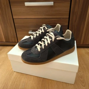 Svarta maison Martin margiela  - Snygga svarta maison Martin margiela sneakers med beige snörning och gummisula. Ovandelen är i en mix av mocka och skinn, vilket ger en stilren och sportig look. Perfekta för dig som gillar klassisk design med moderna detaljer.