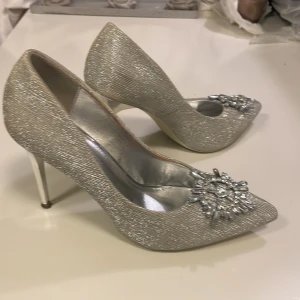 Glittriga silverpumps med strassdetalj - Snygga pumps i glittrigt silver med spetsig tå och hög klack. Skorna har en dekorativ strassdetalj framtill som ger extra bling. Perfekta för dig som vill sticka ut och glänsa. Insidan är metallic och utsidan skimrar i silver. skorna passar troligtvis även dig som har storlek 39.