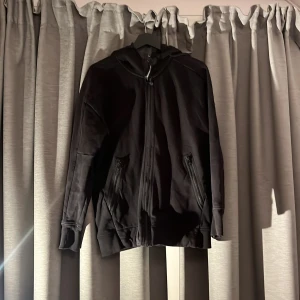 Svart hoodie med goggles från C.P. Company - Svart hoodie från C.P. Company med hel dragkedja, två fickor med dragkedja och ikoniska goggles på huvan. Tröjan har lång ärm och ribbade muddar. Nypris: 3150kr