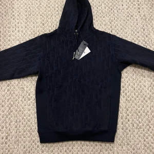 Mörkblå hoodie från D1or - Snygg mörkblå hoodie från Les Deux med diskret mönster i tyget och klassisk huva med snörning. Tröjan har ribbade muddar och en mjuk känsla, perfekt för en chill och stilren look.