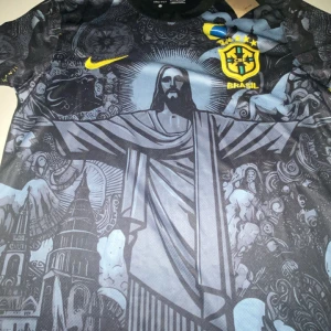 Brasil Nike fotbollströja svart/gul - Unik Brasilien fotbollströja från Nike med svart och grått konstnärligt tryck av Kristusstatyn och brasilianska motiv. Gult Nike-märke och Brasilien-emblem med stjärnor på bröstet. Tillverkad i lätt, ventilerande material. Perfekt för dig som vill sticka ut på planen.
